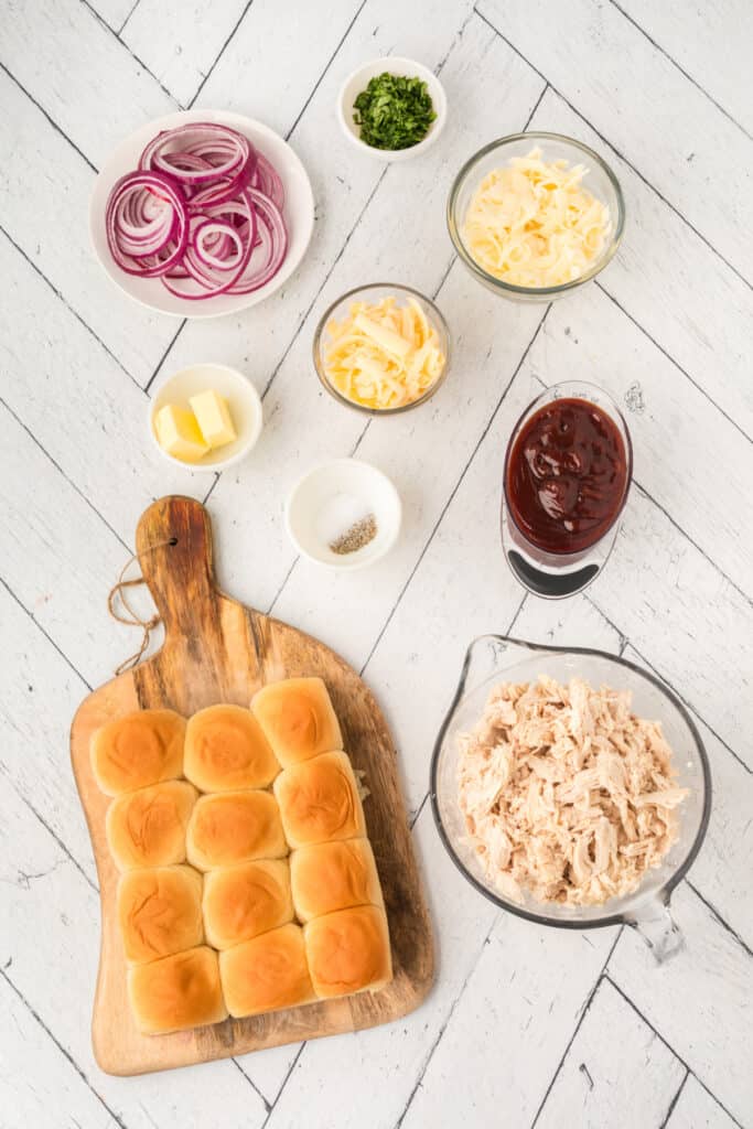 Ingredients for BBQ pulled chicken sliders including slider buns, shredded chicken, BBQ sauce, cheeses, red onion, cilantro, and butter.