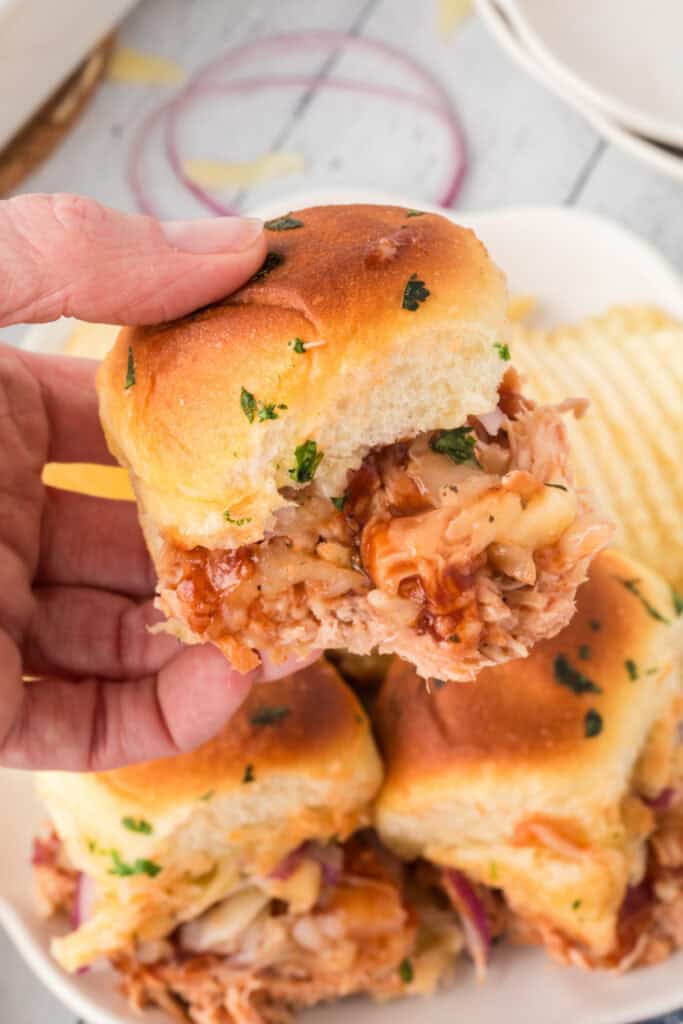 Hand holding a cheesy BBQ pulled chicken slider with red onion and cilantro.