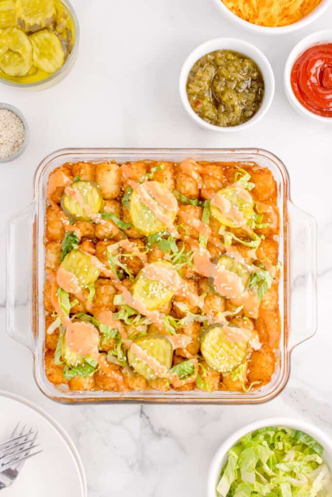 Baked Big Mac Tater Tot Casserole topped with pickles, shredded lettuce, Thousand Island dressing, and sesame seeds.