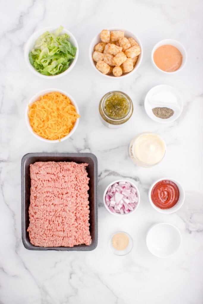 Ingredients for Big Mac Tater Tot Casserole arranged in bowls with ground beef, tater tots, cheese, lettuce, relish, sauces, onion, and seasonings.