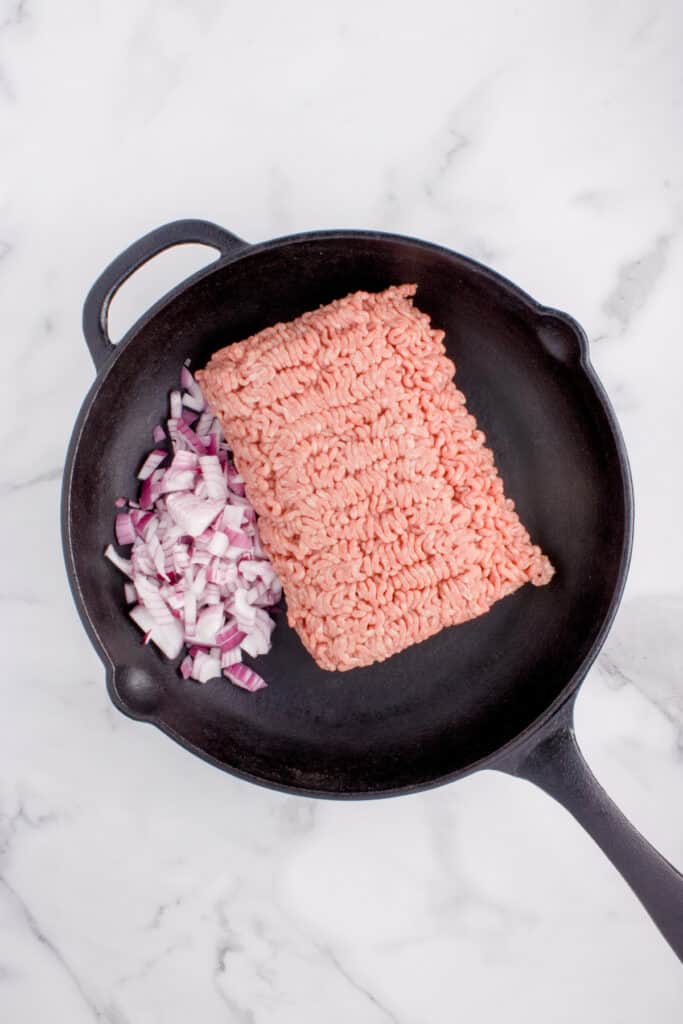 Raw ground beef and diced onion in a cast-iron skillet for Big Mac Tater Tot Casserole.